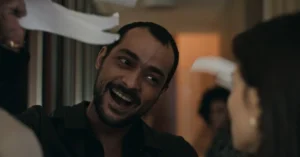Sérgio, interpretado por Eduardo Sterblitch, sorri de forma provocativa enquanto segura papéis rasgados durante uma discussão no corredor do condomínio.