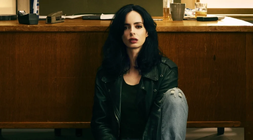 Jessica Jones sentada no chão encostada em uma mesa de madeira, com expressão séria