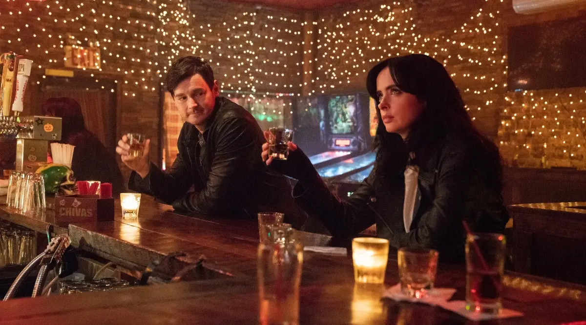 Jessica Jones sentada em um bar com outro personagem, cena noturna iluminada com luzes