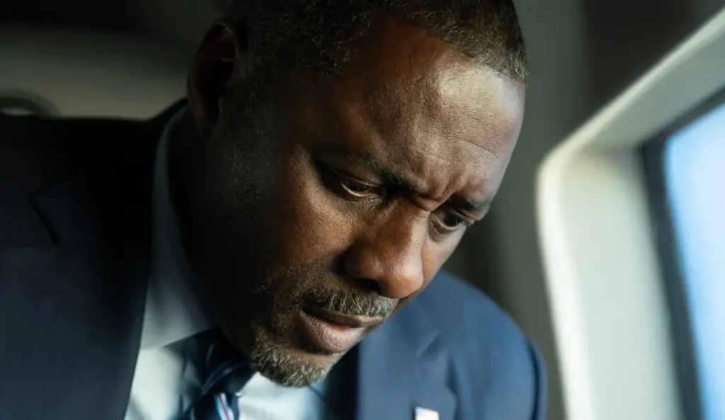 Idris Elba em close, usando terno azul e gravata, demonstrando tensão em cena do filme Casa de Dinamite (2025).