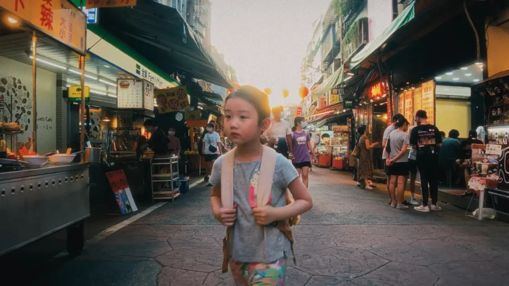 A menina I-Jing atravessando um mercado iluminado enquanto carrega sua mochila no filme A Garota Canhota.