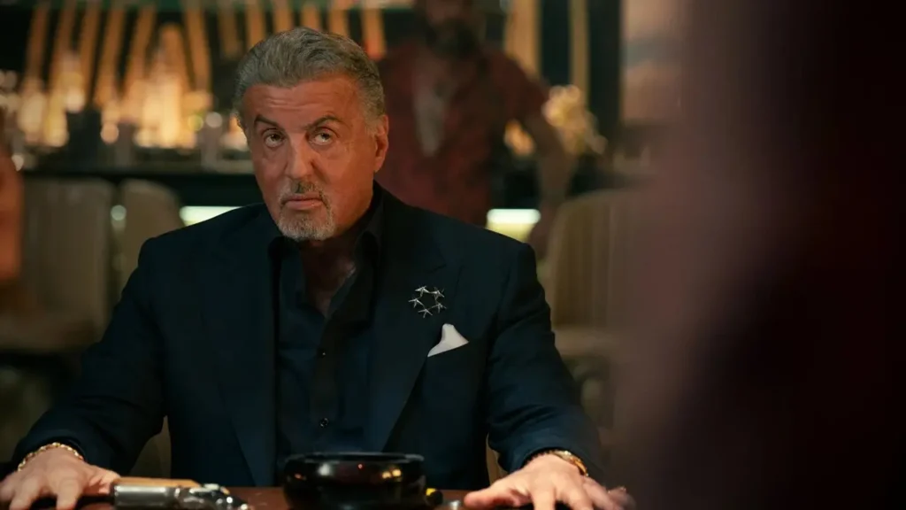 Sylvester Stallone como Dwight Manfredi sentado à mesa em uma sala escura na terceira temporada de Tulsa King