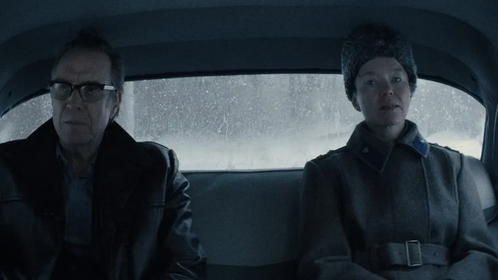 Dois personagens de Cidade das Estrelas aparecem sentados no banco traseiro de um carro, em uma cena com clima frio e sombrio