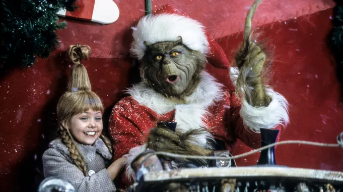 O Grinch vestido de Papai Noel segura as rédeas de um trenó com expressão de surpresa, ao lado da sorridente menina Cindy Lou Who sob a neve.